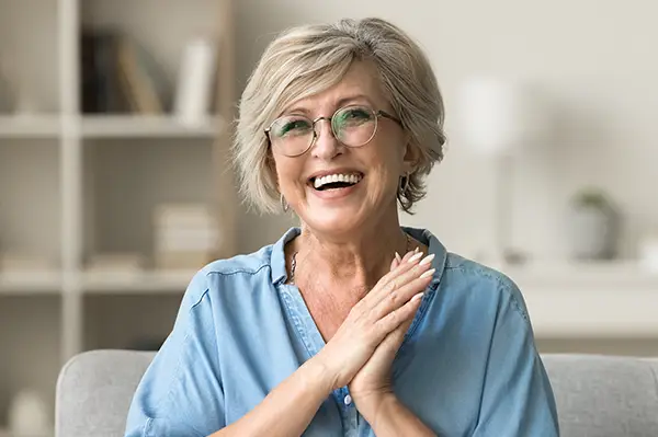 Cheerful pretty older woman in elegant glasses sitting on cozy home couch with dental implants at Fairbanks Periodontal Associates in Fairbanks, AK