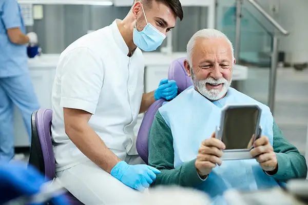 Happy, senior patient looking at himself in a mirror with dental implants at Fairbanks Periodontal Associates in Fairbanks, AK